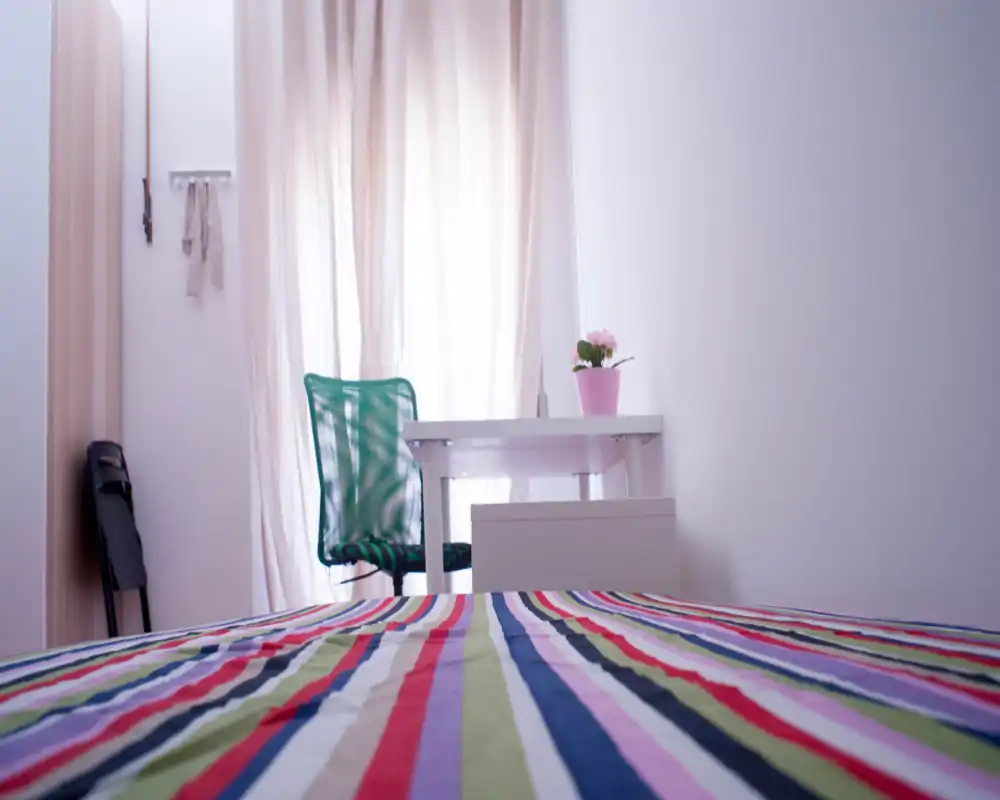 Bright, modern bedroom view with a colorful striped bed in the foreground, a simple desk and green chair by a sheer curtained window, and a small potted plant adding a cozy touch.