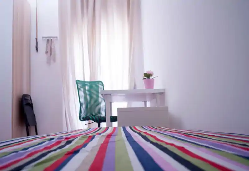 Bright, minimalist bedroom view with a colorful striped bedspread, a simple desk by the window and a green mesh chair — ideal for a single occupant or student.