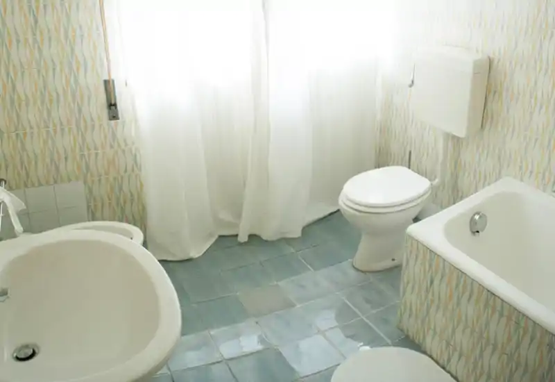 Bagno luminoso piastrellato con vasca, WC e lavabo; luce naturale diffusa da una finestra con tenda.