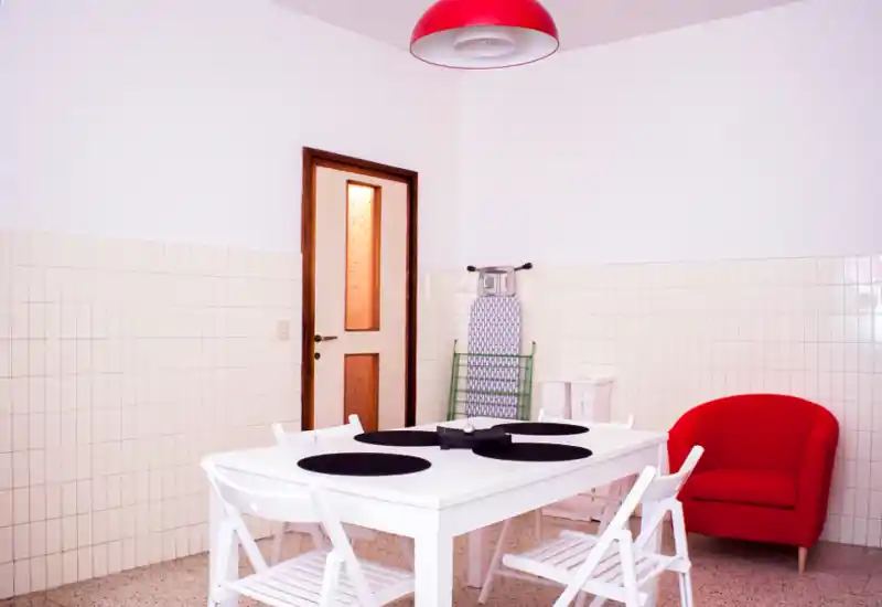 Zona pranzo luminosa e minimal con tavolo bianco e sedie pieghevoli, poltrona rossa di design e arredi essenziali — perfetta per pasti accoglienti.