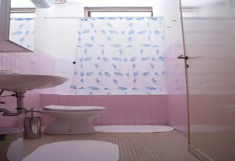 Bagno compatto e luminoso con piastrelle rosa, vasca con tenda decorata, lavabo e WC — pulito e dallo stile semplice.