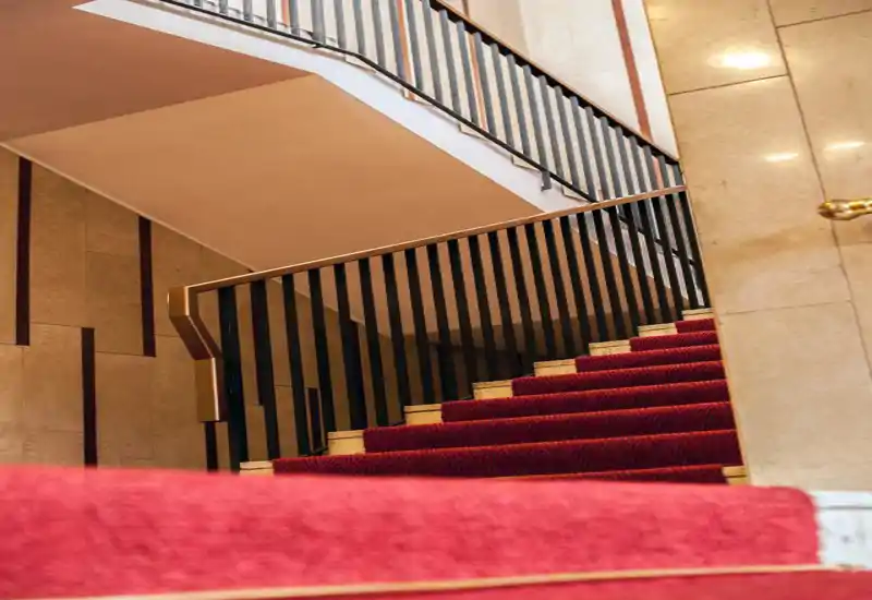 Scalinata d'ingresso con passamaneria rossa vellutata e ringhiera a stecche metalliche; pareti marmoree e illuminazione calda creano un'atmosfera elegante.