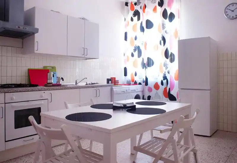 Cucina luminosa e moderna con pensili bianchi, tavolo con sedie, elettrodomestici (forno e frigorifero) e tende colorate che lasciano entrare luce naturale.
