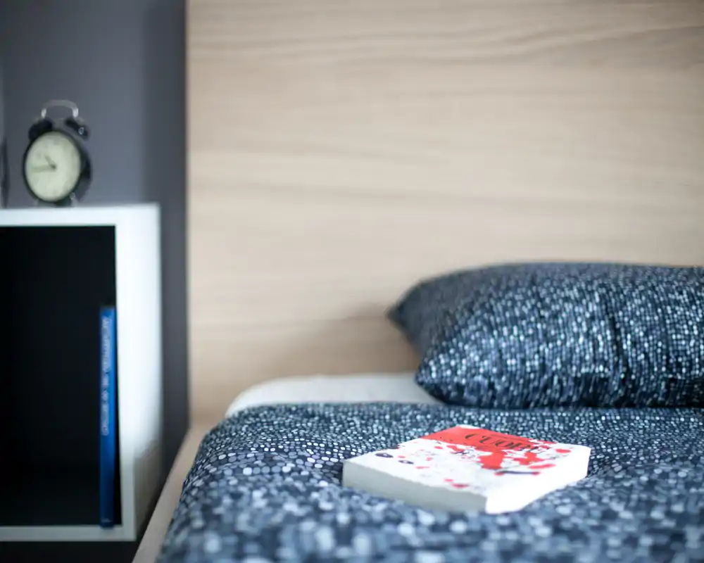 Gros plan d'une chambre soignée avec un lit confortable, linge à motifs, un oreiller et une table de chevet avec réveil — lumineux et accueillant, parfait pour une annonce.