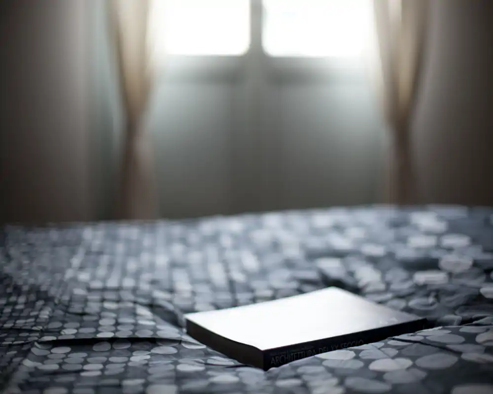 Scène de chambre minimaliste avec une couette à motifs et un livre posé sur le lit, éclairée par une fenêtre en contre‑jour apportant une lumière naturelle douce.