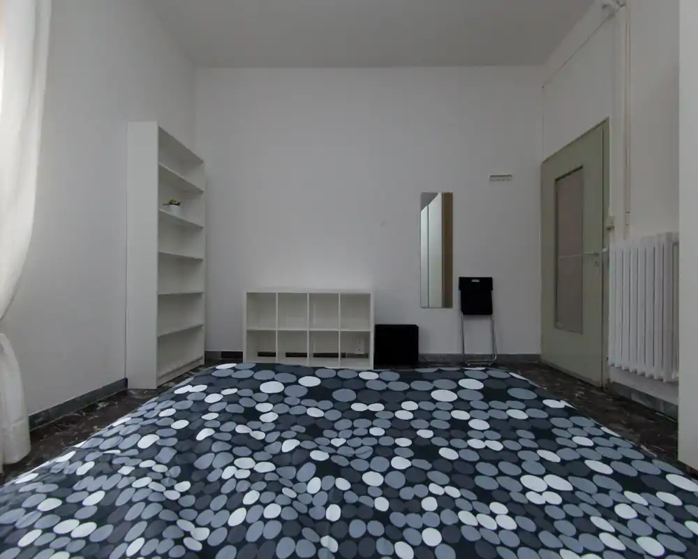 Chambre minimaliste et lumineuse avec couette à motifs au premier plan, murs blancs, étagères et miroir mural. Style épuré et lumière naturelle venant de la gauche.