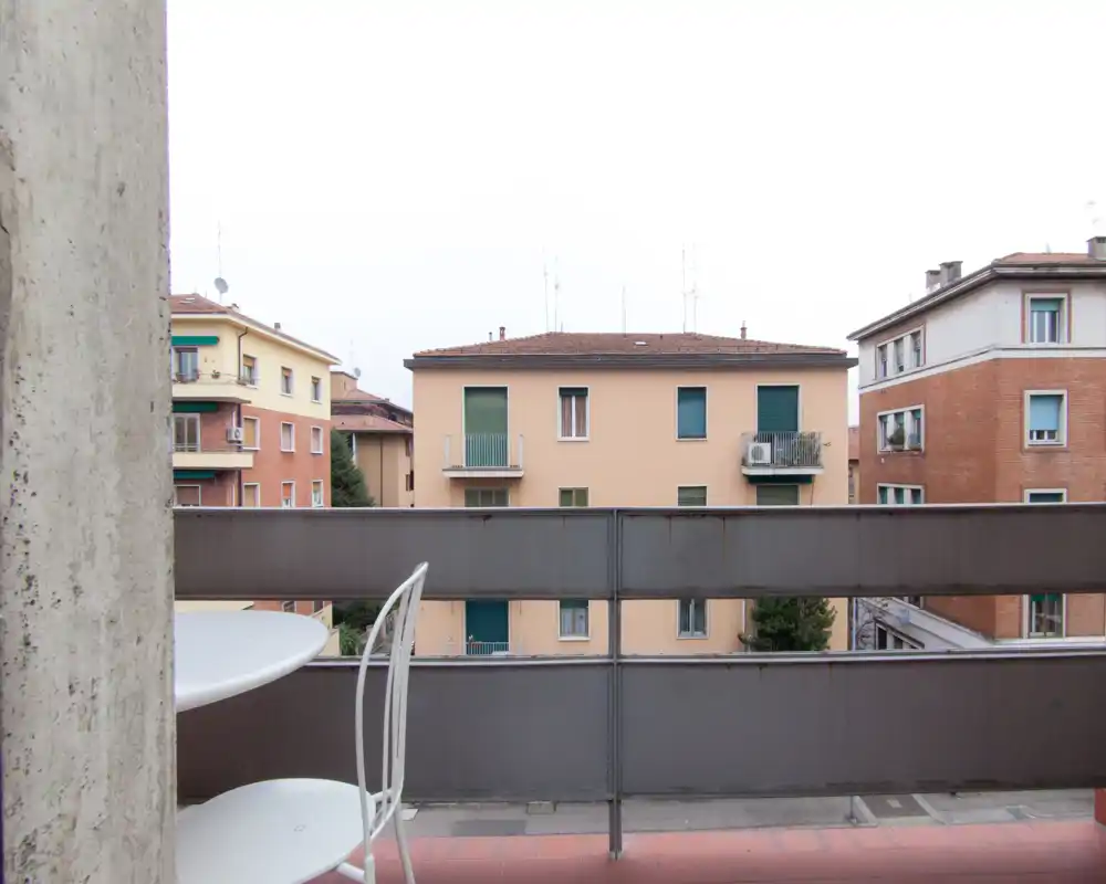 Petit balcon meublé avec garde-corps métallique et une table avec chaise, donnant sur des immeubles voisins — parfait pour un café du matin.