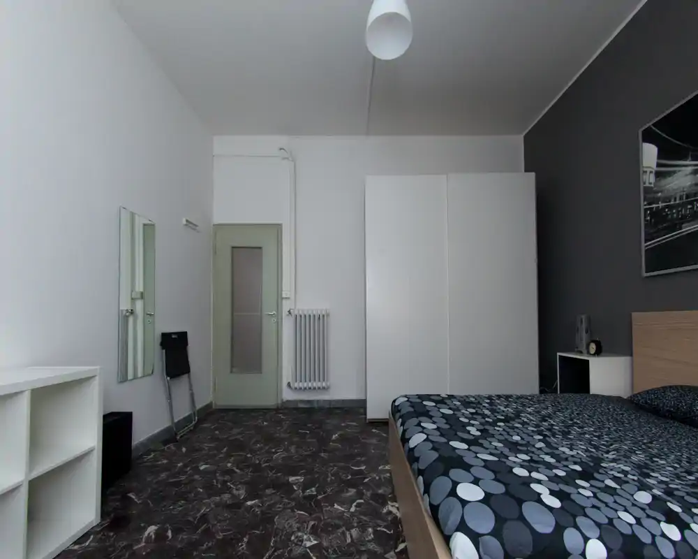 Chambre moderne et minimaliste avec un lit double au premier plan à droite, une grande armoire blanche et une table de chevet sobre ; décoration neutre et linge de lit à motifs pour un rendu propre et fonctionnel.