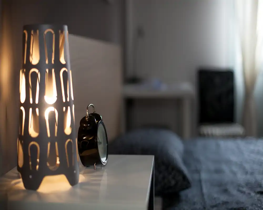 Coin chambre chaleureux avec une lampe de chevet allumée et un réveil sur une table de nuit blanche ; le lit et les textiles doux sont visibles en arrière-plan avec un éclairage chaud et accueillant.
