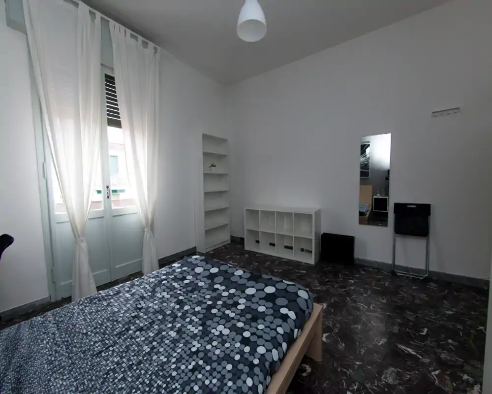 Chambre minimaliste et lumineuse avec lit double, couette à motifs, grande fenêtre aux rideaux blancs et étagères basses pour rangement — espace calme et dégagé adapté à la location.