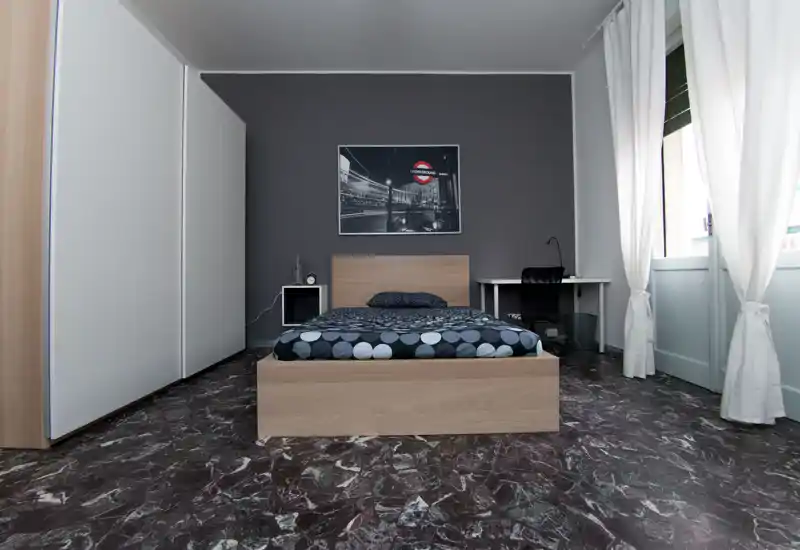 Dormitorio moderno y minimalista con cama de madera centrada frente a una pared oscura, gran armario, escritorio junto a la ventana y ropa de cama con estampado. La luz natural entra a través de las cortinas blancas, creando un espacio limpio y bien compuesto, perfecto para anuncios.