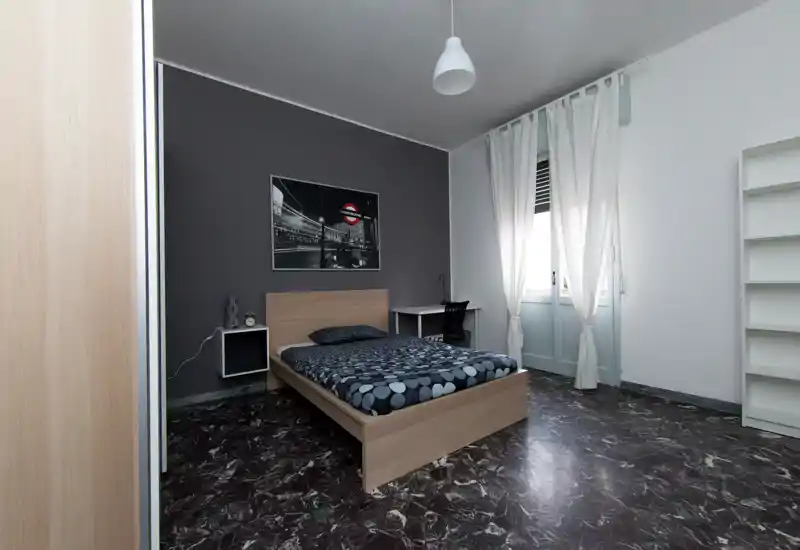 Dormitorio luminoso y de mobiliario sencillo con cama doble, estante de mesilla, escritorio junto a la ventana y decoración neutra. La luz natural entra por las cortinas blancas y baña el suelo de mármol con diseño.
