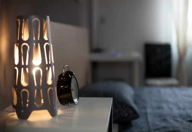 Detalle cálido e íntimo de un dormitorio con una lámpara encendida y un despertador en la mesita, con la cama desenfocada al fondo que aporta una atmósfera acogedora.