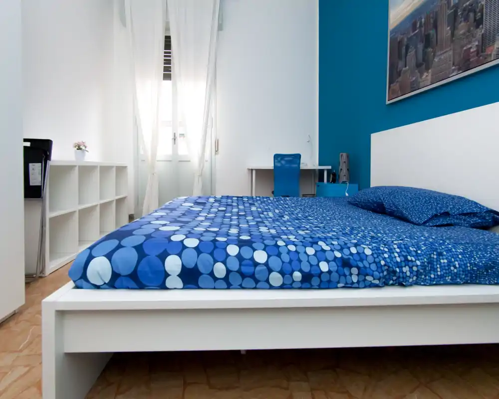 Chambre moderne et lumineuse avec un grand lit blanc et une literie bleue à motifs. Style minimaliste soigné, lumière naturelle par la fenêtre et un petit bureau à l'arrière-plan.