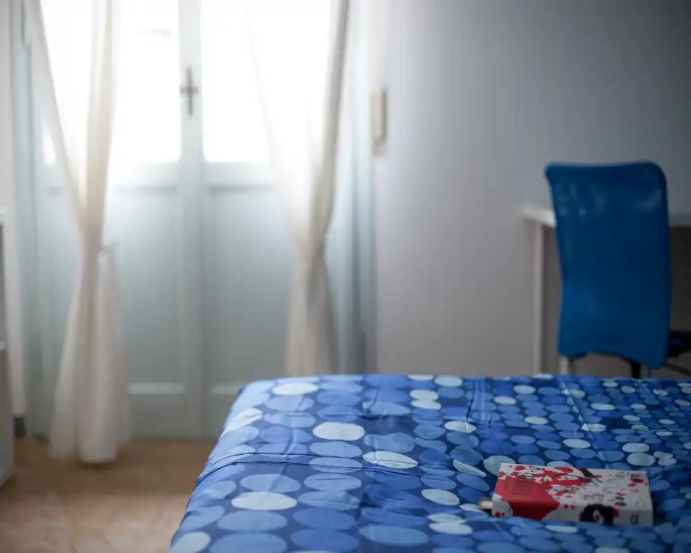 Chambre lumineuse au style simple avec une couette bleue à motifs au premier plan et une lumière naturelle douce provenant de la fenêtre aux rideaux clairs.