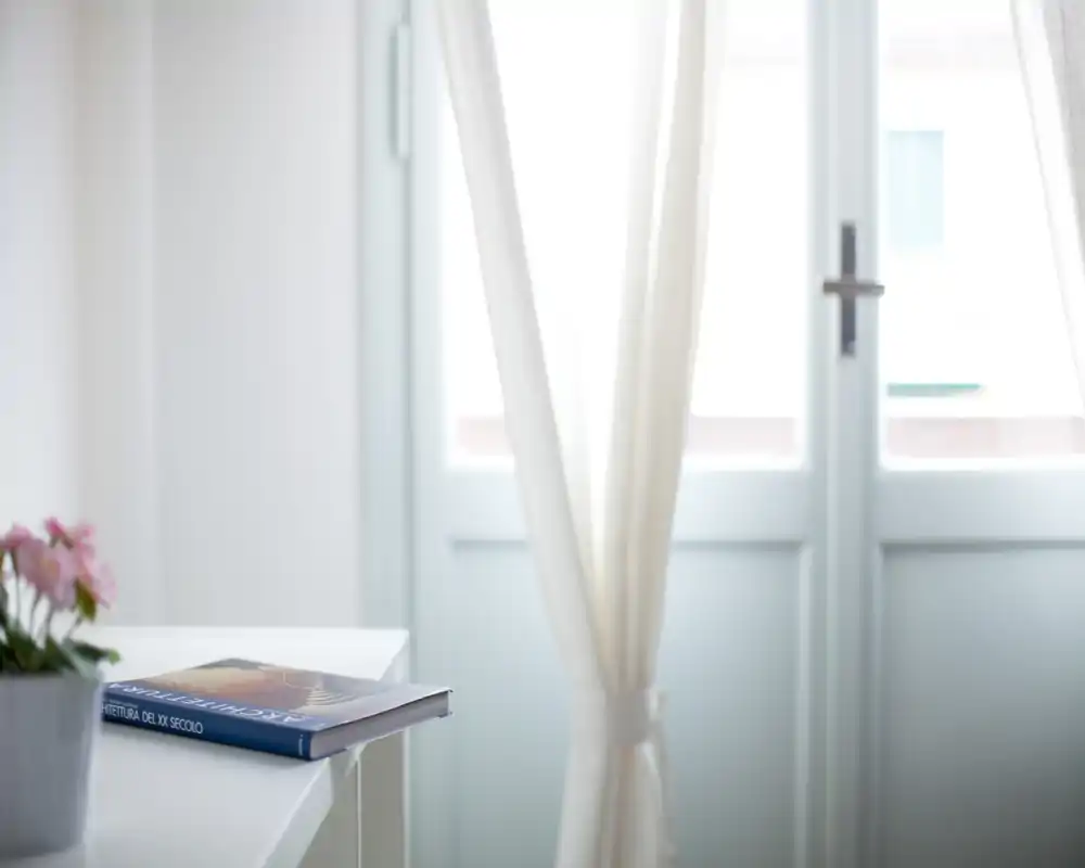 Espace séjour lumineux et minimaliste avec des voilages et une petite table portant un livre et une plante en pot, mettant en valeur la lumière naturelle.