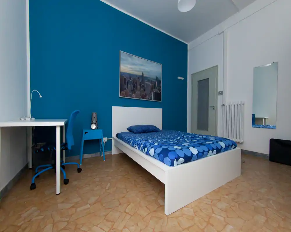 Chambre simple lumineuse et bien mise en scène avec lit blanc, literie bleue assortie et coin bureau — propre et fonctionnelle pour la location courte durée.