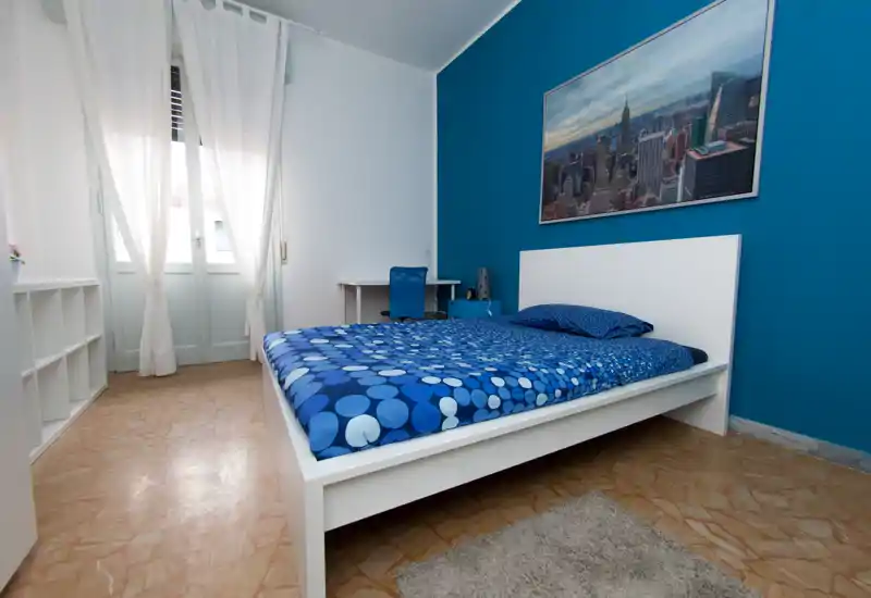 Chambre lumineuse et bien mise en scène avec un lit à plateforme blanc et un mur bleu. La lumière naturelle provenant de la fenêtre met en valeur la literie propre et le mobilier moderne et épuré.