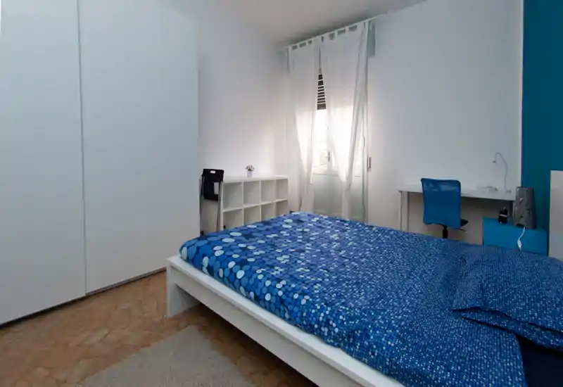 Chambre lumineuse et soignée avec un grand lit à housse bleue à motifs, une armoire blanche, bureau et chaise près de la fenêtre ; espace minimaliste et bien éclairé, idéal pour un séjour court.