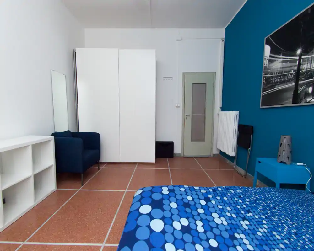 Chambre moderne et lumineuse aux accents bleus, lit double visible avec couette à motifs, armoire et fauteuil confortable.