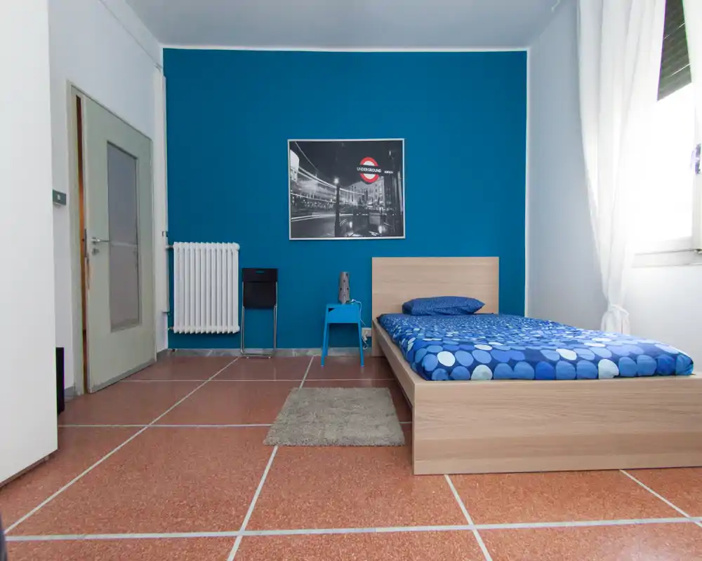 Chambre simple et lumineuse avec un lit en bois, un mur bleu accentué et une lumière naturelle provenant de la fenêtre. Mobilier minimaliste avec table de chevet, radiateur et cadre au mur.
