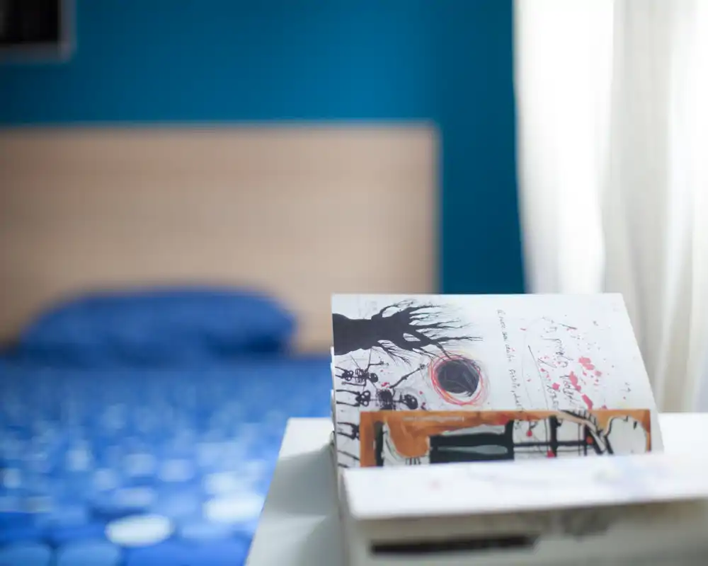 Chambre lumineuse et douce avec une housse de lit bleue et une tête de lit en bois clair ; profondeur de champ faible mettant en valeur des cartes illustrées sur la table de chevet.