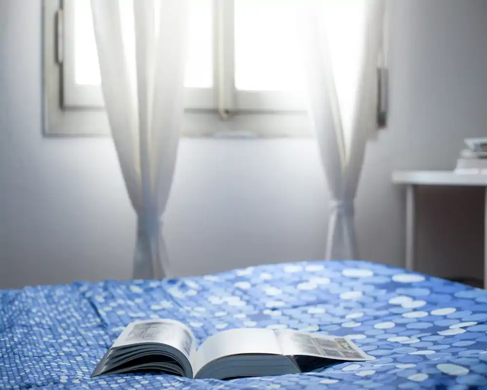 Chambre lumineuse et sobre avec une couette bleue à motifs et un livre ouvert sur le lit; une lumière naturelle douce filtre à travers les voilages, créant une ambiance apaisante.