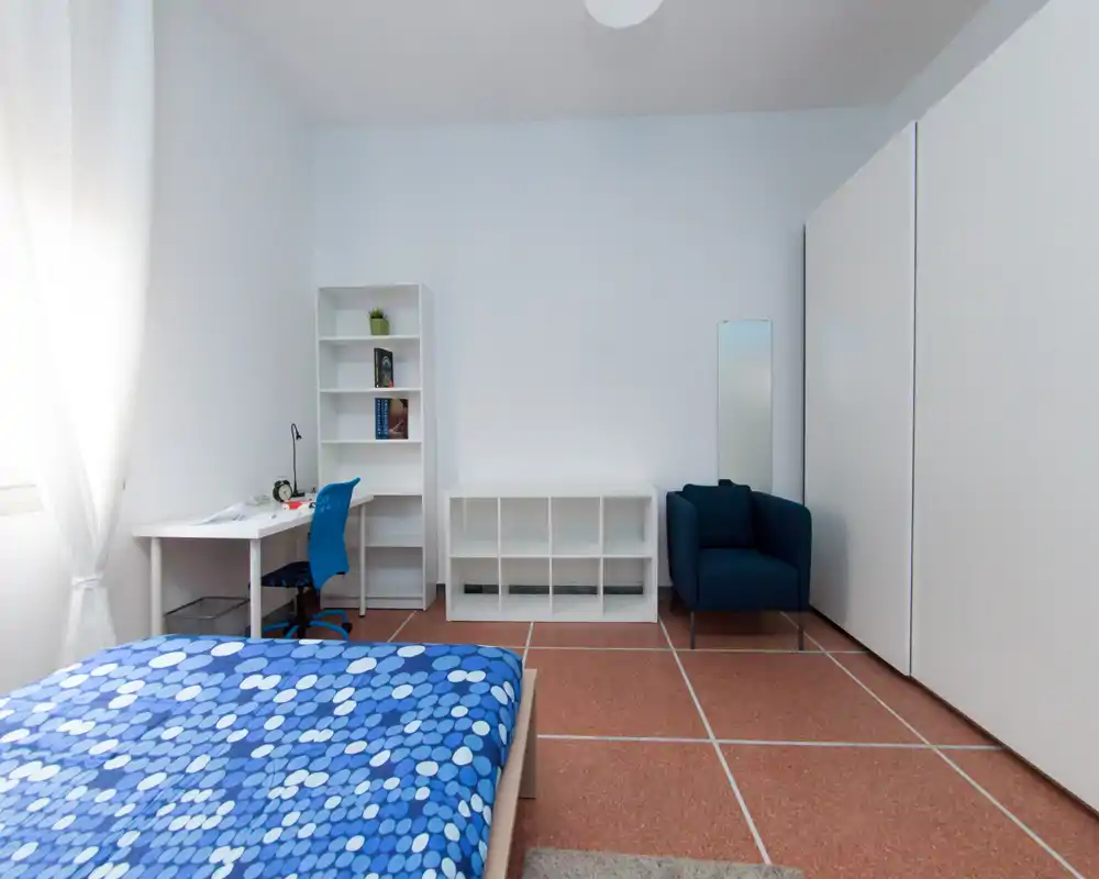 Chambre lumineuse et minimaliste avec un lit double en couvre-lit bleu à motifs, un bureau près de la fenêtre, des étagères et un fauteuil confortable — bien éclairée et sobrement meublée.
