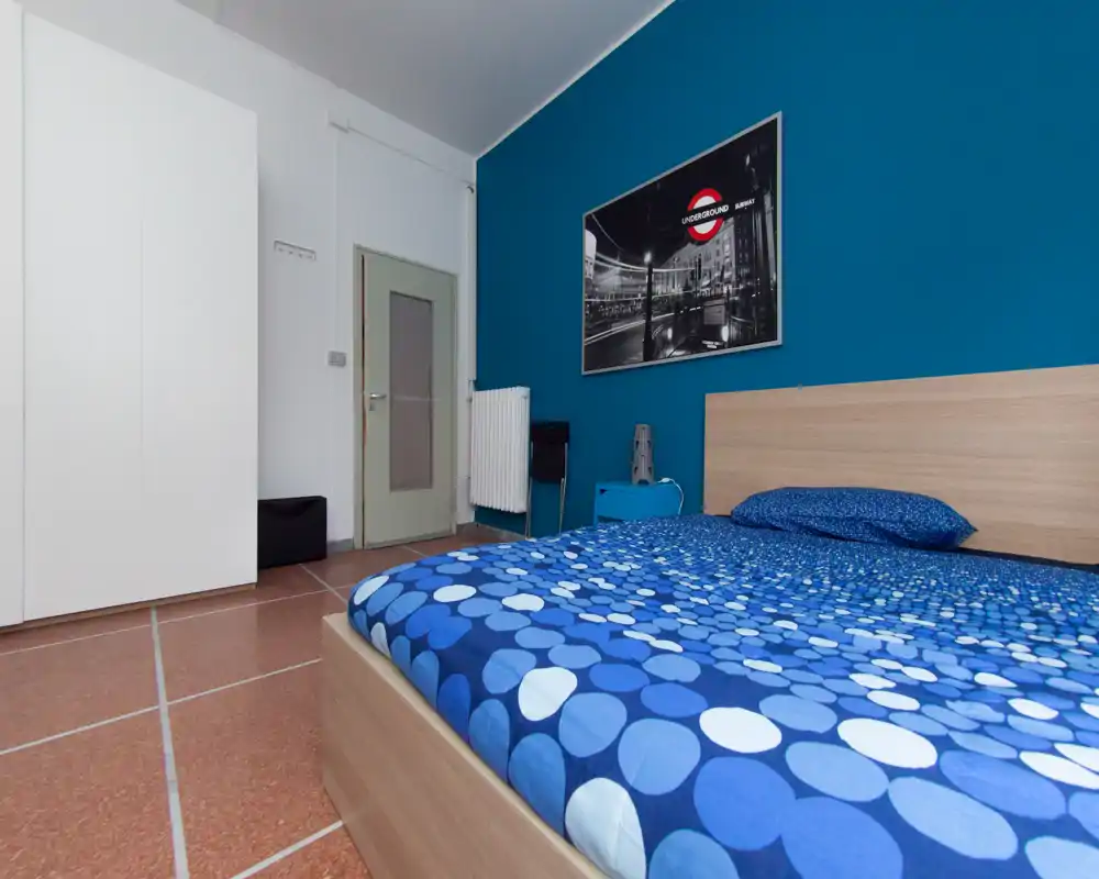 Chambre moderne et lumineuse avec un lit double en linge bleu à motifs, tête de lit en bois, mur d'accent turquoise et armoire blanche — propre et contemporaine.