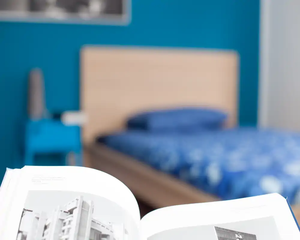 Photo lumineuse d'une chambre moderne avec un lit en bois et une literie bleue; un livre ouvert au premier plan apporte une touche lifestyle.