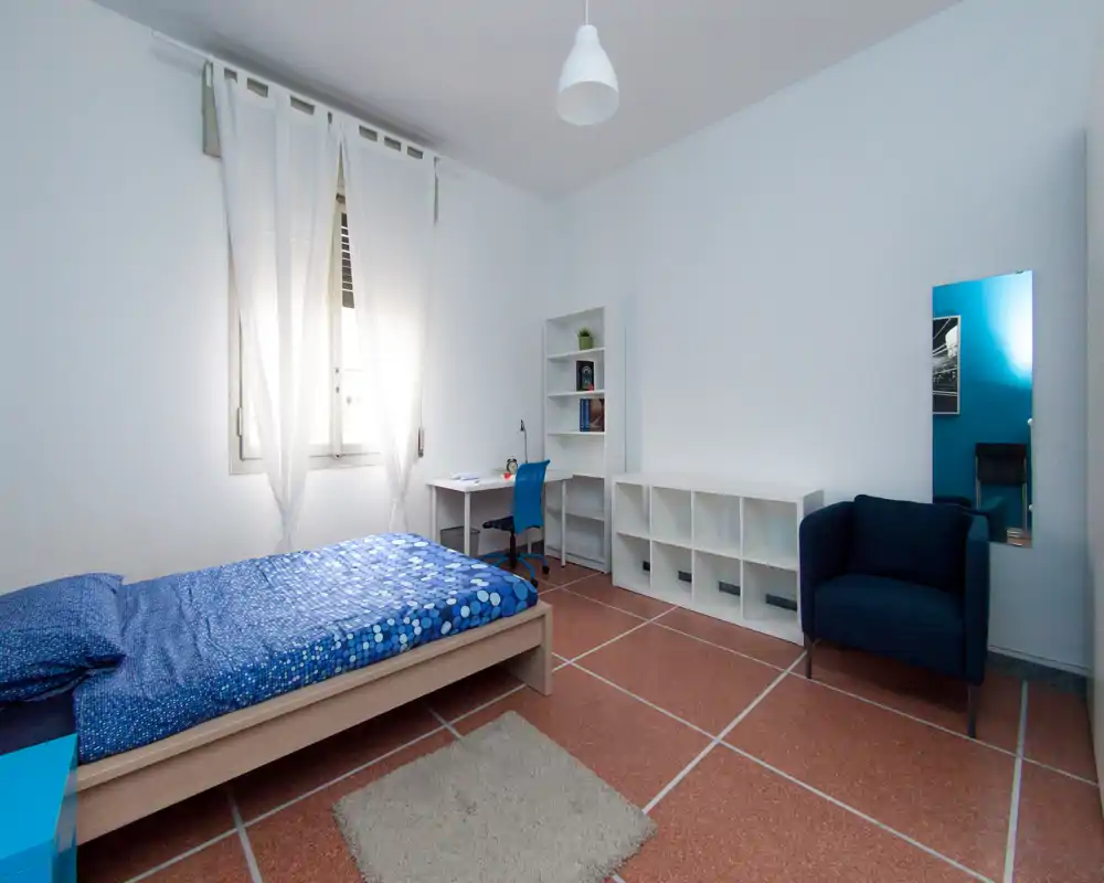 Chambre lumineuse et sobrement meublée avec un lit simple, un bureau et des étagères—idéale pour un étudiant ou une personne seule. La lumière naturelle et les touches de bleu apportent une ambiance paisible.