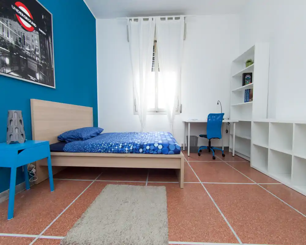 Chambre simple lumineuse et moderne avec lit bas en bois, mur d'accent bleu et literie assortie. La pièce comprend un bureau et des étagères, idéale pour un étudiant ou une personne seule.
