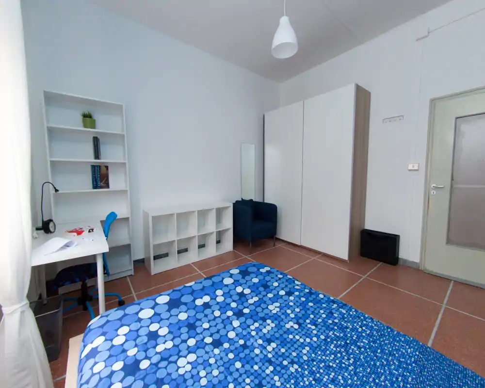 Chambre lumineuse et soignée avec lit double recouvert d'une couette bleue à motifs, bureau près de la fenêtre, étagère et armoire — idéale pour un étudiant ou une personne seule.