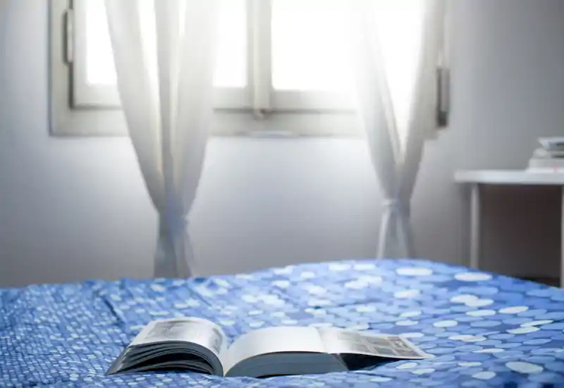 Camera da letto luminosa e accogliente con copriletto blu a motivi e un libro aperto sul letto; la luce naturale soffusa dalla finestra con tende crea un’atmosfera tranquilla.