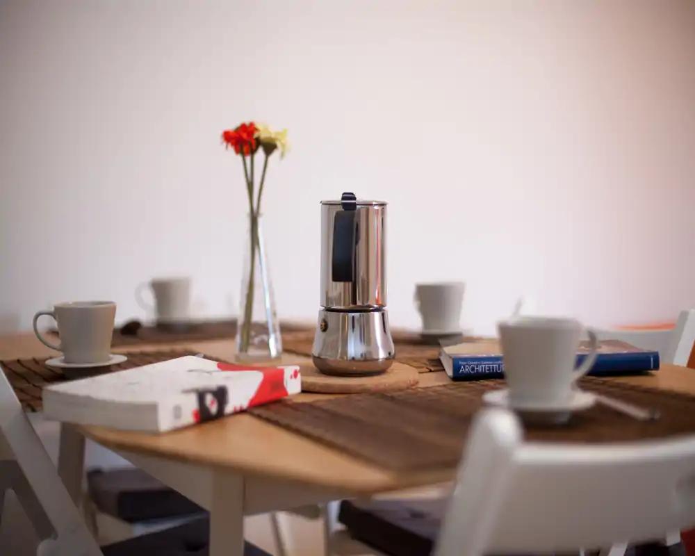 Table à manger décorée avec tasses, sets de table, une cafetière moka et un petit vase — cadre lumineux et accueillant, idéal pour petit-déjeuner ou café.