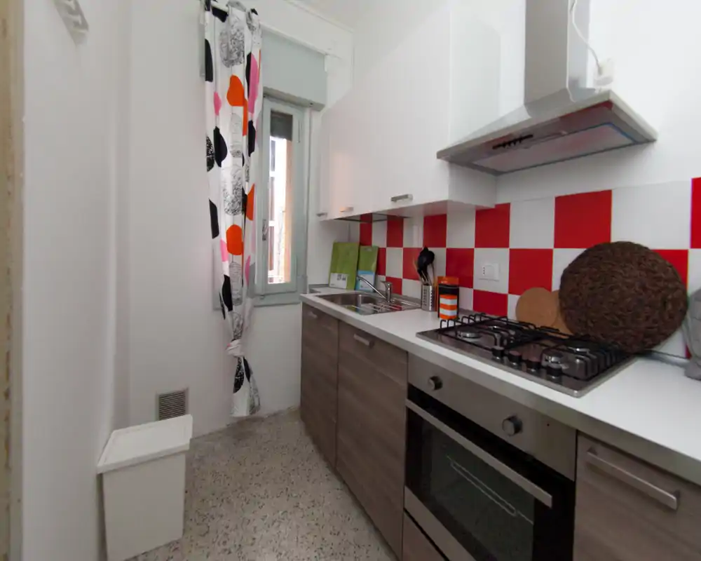 Cuisine moderne et compacte avec meubles bas effet bois, placards blancs et crédence à carreaux rouge et blanc. Équipée d'une plaque à gaz, d'un four encastré, d'un évier inox et d'une fenêtre lumineuse apportant de la lumière naturelle.
