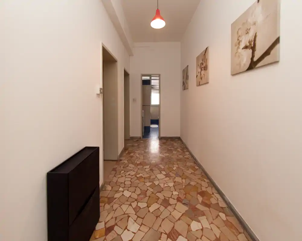Long couloir d'entrée bien éclairé avec sol en terrazzo à motifs, tableaux simples aux murs et suspension lumineuse — accès propre et fonctionnel vers les pièces intérieures.