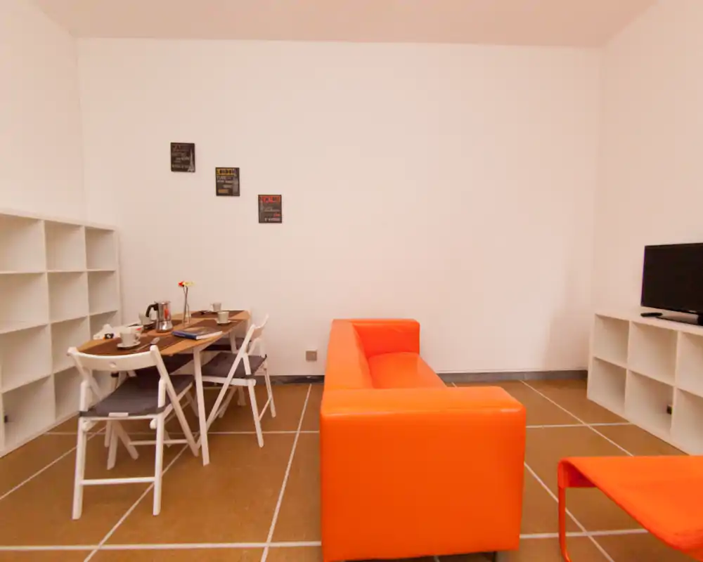 Espace salon/salle à manger soigné avec canapé orange vif, petite table à manger et étagères—mise en scène agréable et lumière douce.