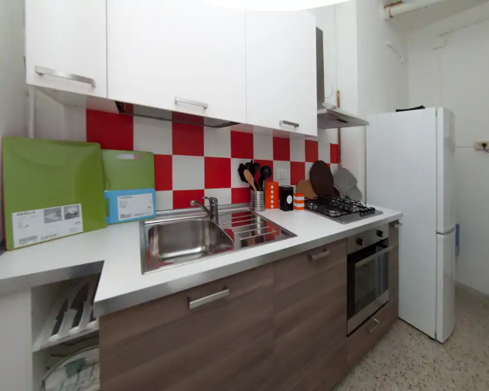 Cuisine compacte et moderne avec évier en inox, plaque à gaz et four encastré, rehaussée d’un crédence à carreaux rouge et blanc et d’un grand réfrigérateur.