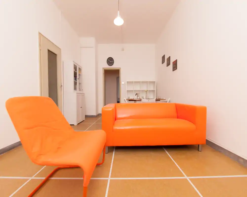 Salon lumineux et minimaliste avec canapé orange vif et fauteuil assorti, murs neutres et petite zone repas/bureau visible à l'arrière-plan.