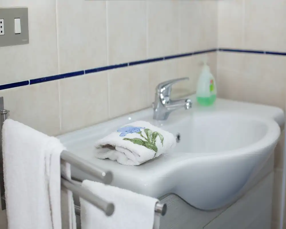 Lavabo de salle de bains propre et bien éclairé avec une serviette décorative pliée et du savon liquide sur le plan, carrelage blanc et robinet chromé.