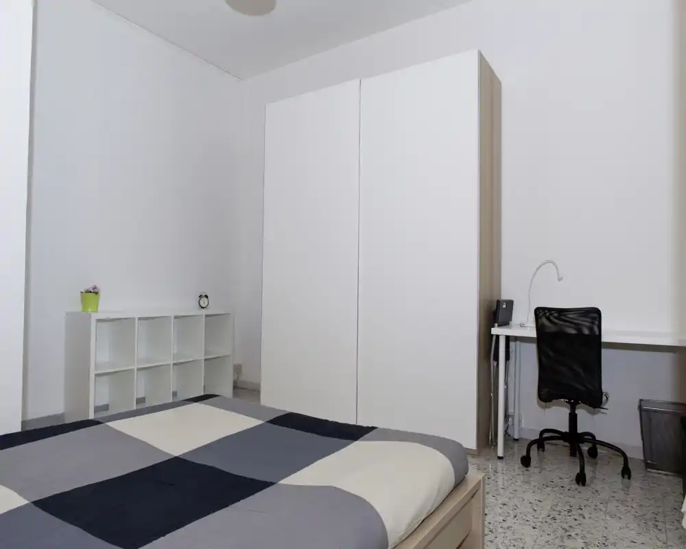 Camera da letto minimal e luminosa con letto matrimoniale con copriletto geometrico, ampia cabina-armadio bianca, scaffalatura e una scrivania compatta con sedia — pulita e moderna.