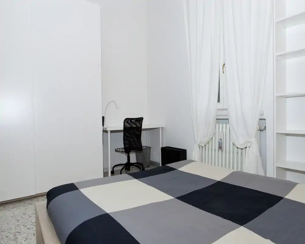 Camera da letto minimal e luminosa con un ampio letto con copriletto a quadri, armadio, scrivania e scaffalature; stile moderno e pulito ideale per annunci.
