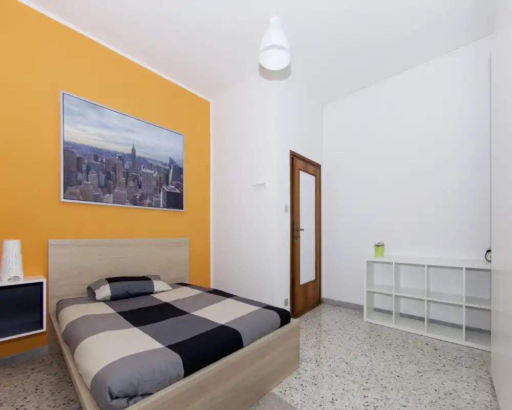 Camera da letto moderna e luminosa con letto matrimoniale contro una parete arancione, arredi essenziali e una grande stampa della città sopra la testata.
