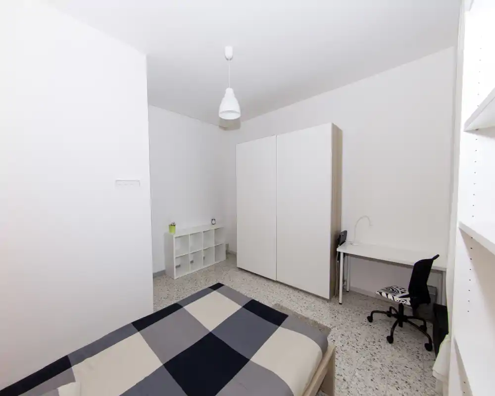 Camera da letto luminosa e minimalista con letto matrimoniale in primo piano, armadio bianco, piccolo scaffale e scrivania con sedia — linee pulite e arredi semplici per uno spazio funzionale adatto agli affitti.