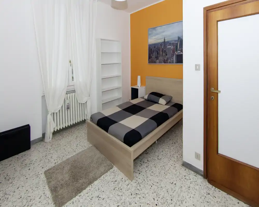 Camera da letto luminosa e arredata in modo semplice con letto singolo su parete arancione, libreria e finestra con tende bianche.