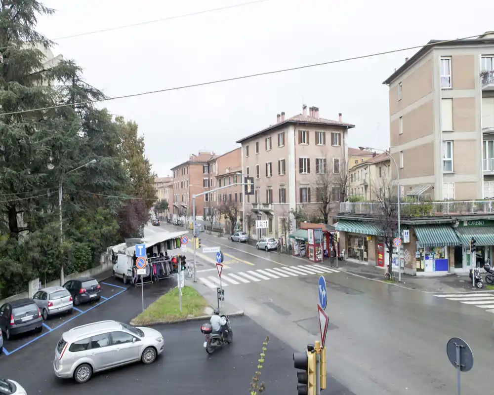 Incrocio urbano con auto parcheggiate, una bancarella e palazzi residenziali; cielo coperto ma vista nitida ideale per mostrare il quartiere.
