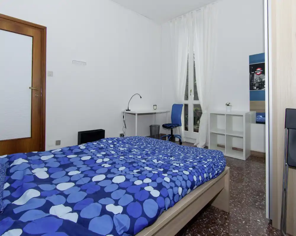 Camera da letto luminosa e semplice con letto matrimoniale con copriletto blu a motivi, piccola scrivania e sedia vicino a una finestra alta che fa entrare luce naturale.