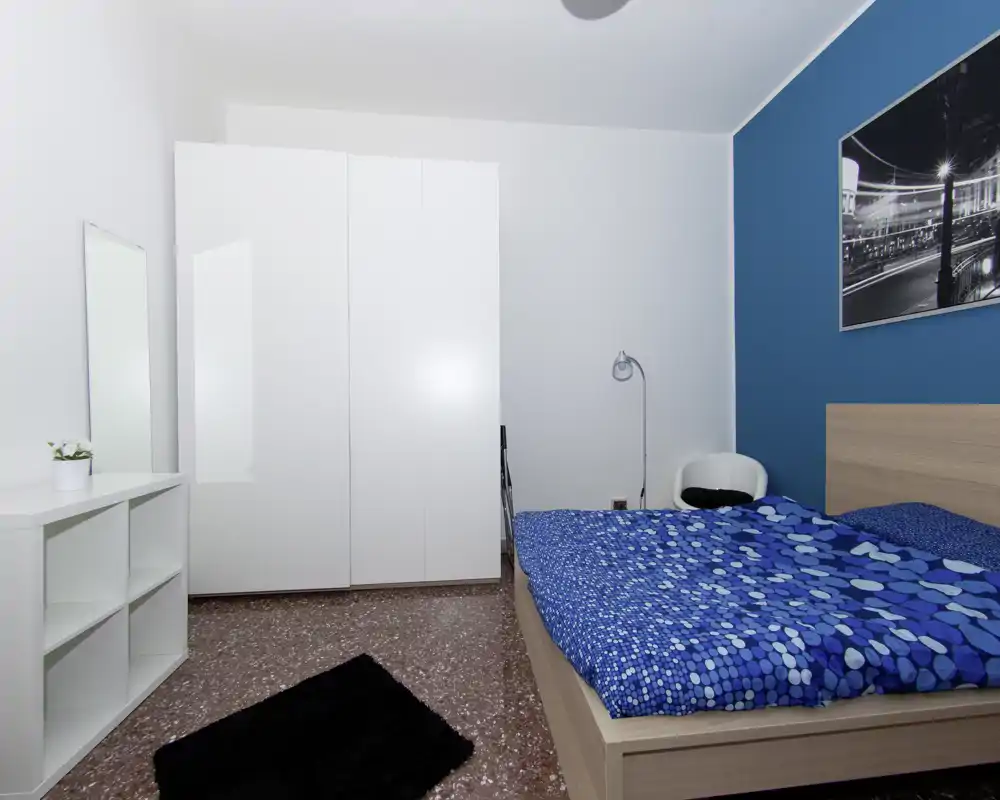 Camera da letto moderna e luminosa con letto matrimoniale, copriletto blu a motivi, armadio bianco e arredi minimali per un ambiente pulito e confortevole.