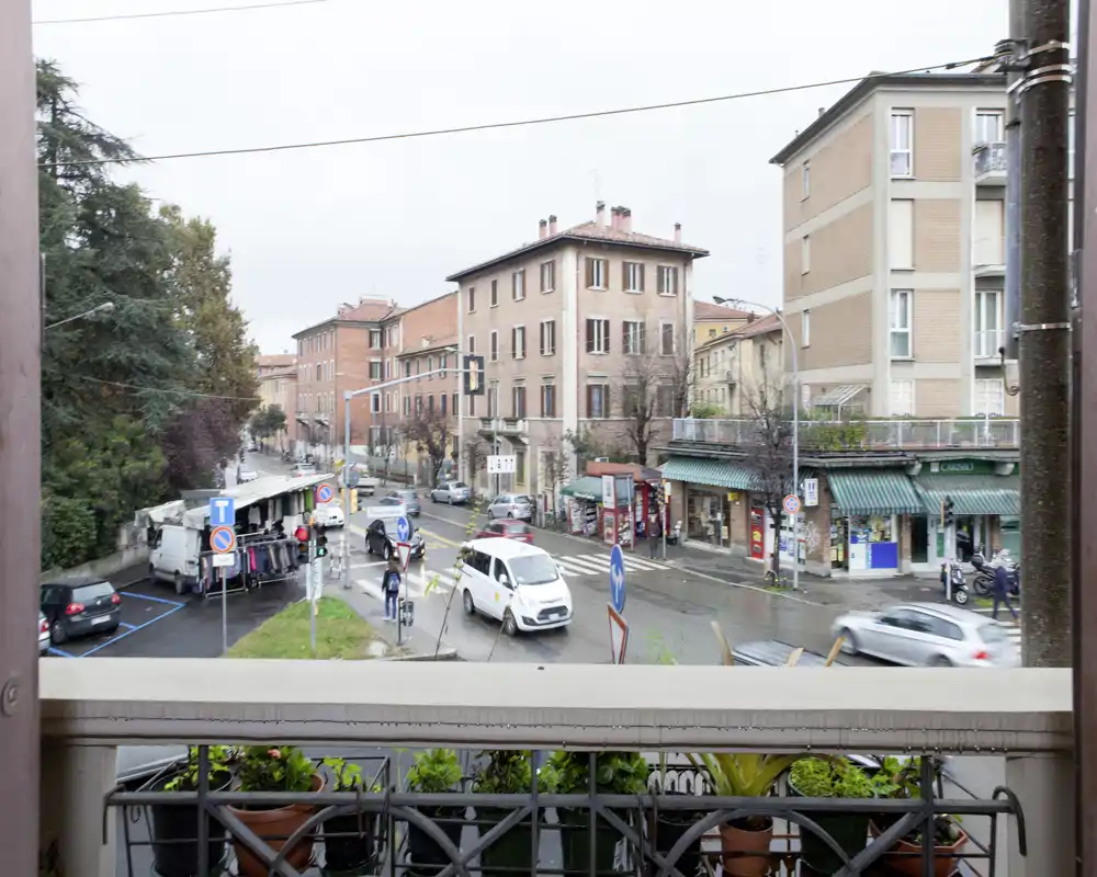 Vista dal balcone su una strada cittadina con negozi e palazzi residenziali; vaso e ringhiera in primo piano incorniciano la scena.